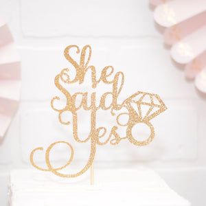 gold she said yes cake topper on a white cake and pink decorations
