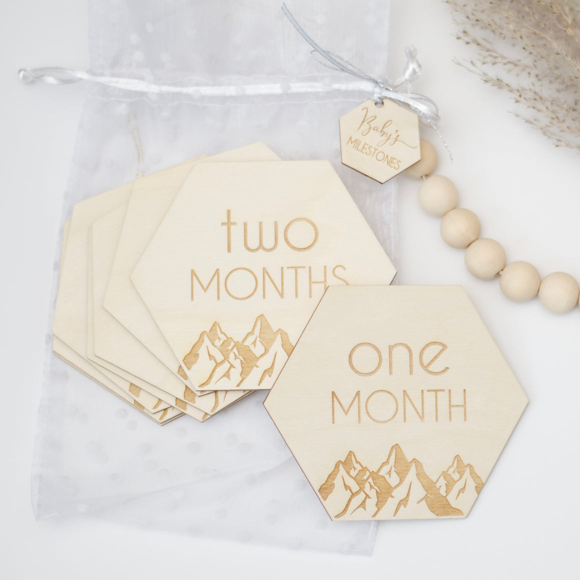 Hexagon shaped wooden milestone signs. One month, two months. Displayed on a white table with wooden beads and plants