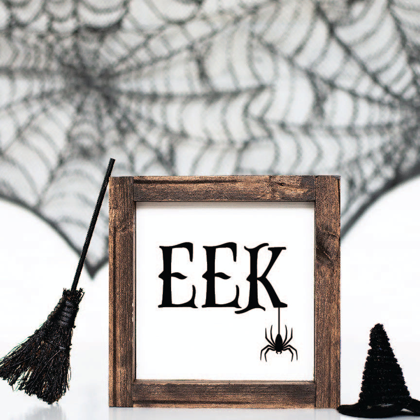 Eek Halloween sign with a spider hanging down from the letters. Sign is framed in a dark brown frame and is styled with mini decorations like a spider web, broomstick and hat. 