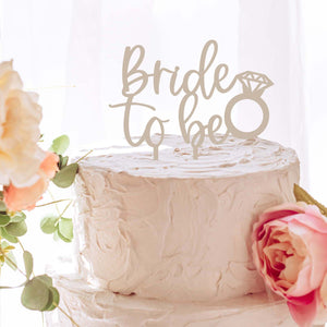 Bride to be cake topper with a diamond ring on top of a white cake with florals