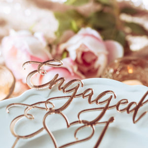 engaged cake topper with heart close up of mirrored rose gold acrylic and pink flowers in the background