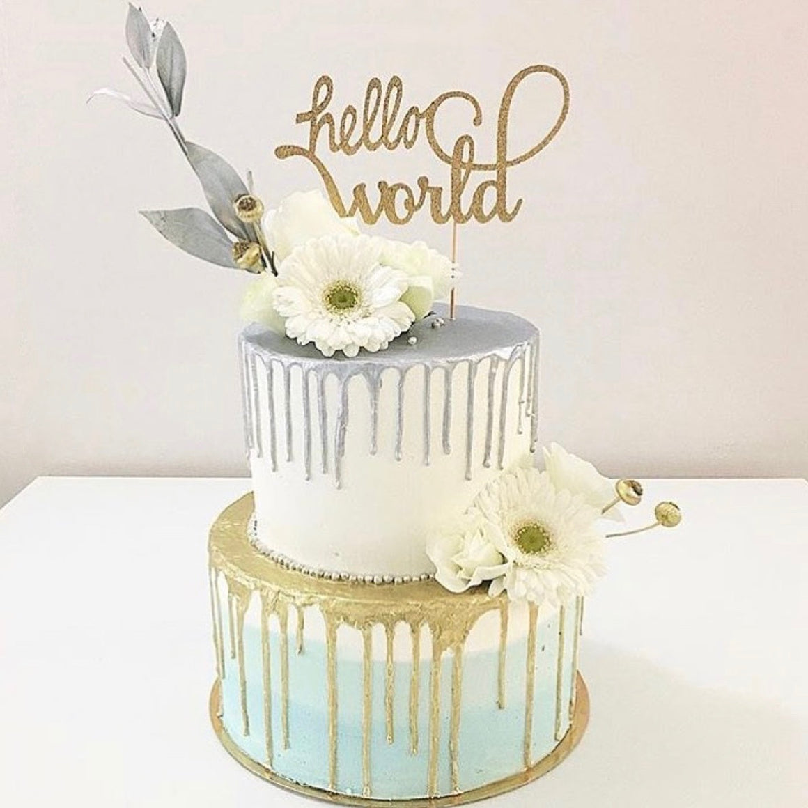 gold hello world cake topper on a silver mint and gold drip cake with daisies