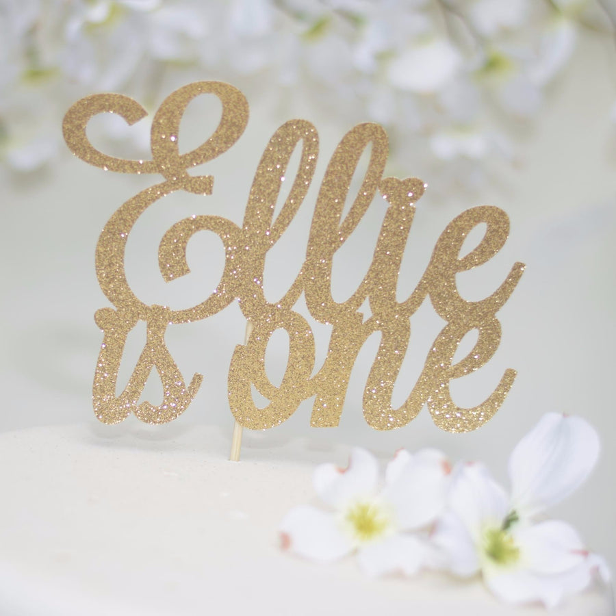 Emma is one gold glitter cake topper on white background wit pink and green felt flower details