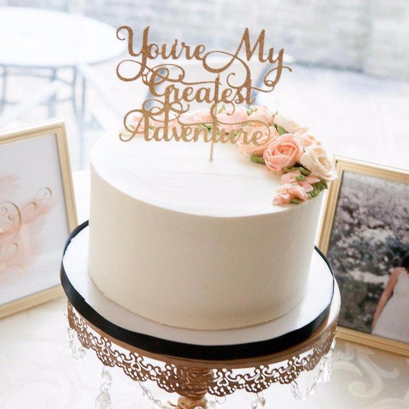 You're my greatest adventure gold glitter cake topper on white wedding cake with a photo of a couple in the background
