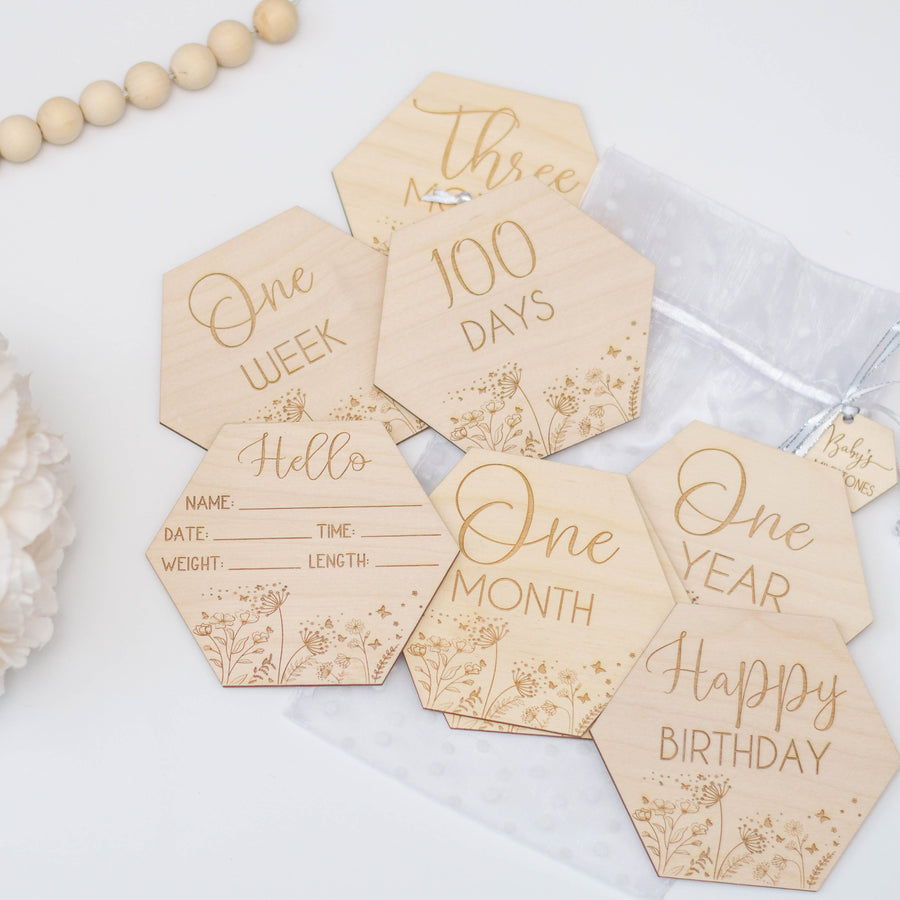 Hexagon shaped baby milestone discs that are engraved onto wood. Displayed on a white table with florals and wooden beads.