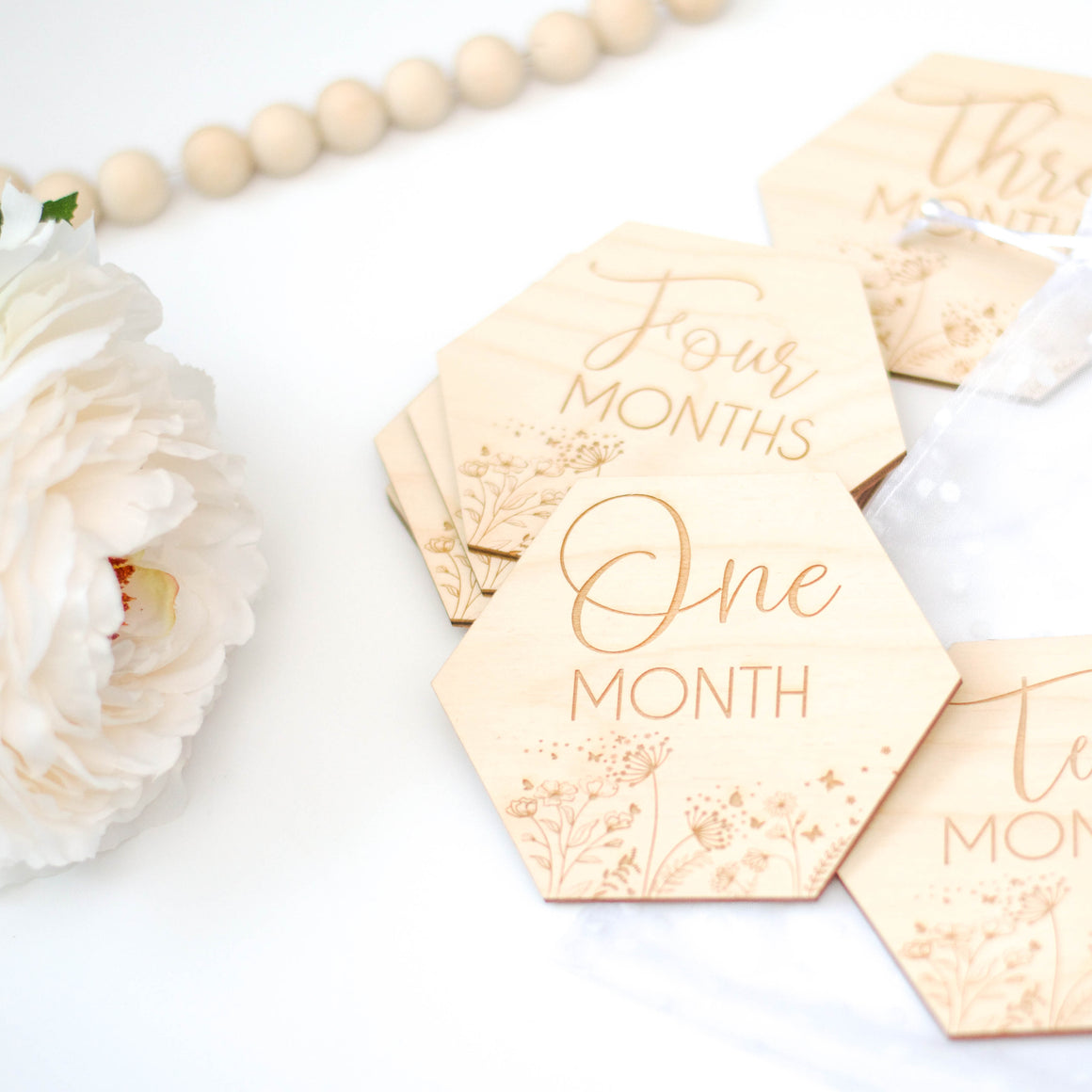 Hexagon shaped baby milestone discs that are engraved onto wood. Displayed on a white table with florals and wooden beads.